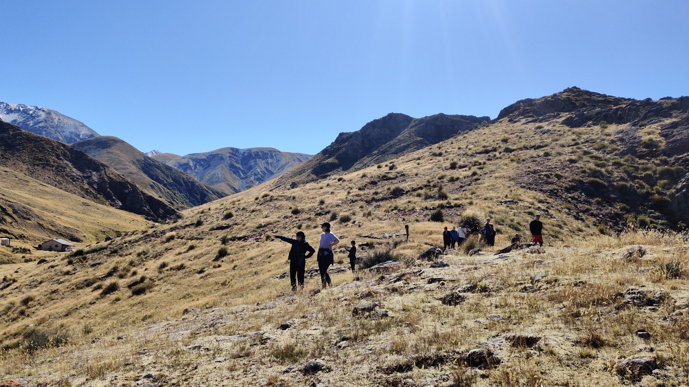 The brown grassy landscape at Woolshed Creek fills the frame, mountain peaks visible in the background. In the midground people are a group of people, small figures, one person is pointing into the distance