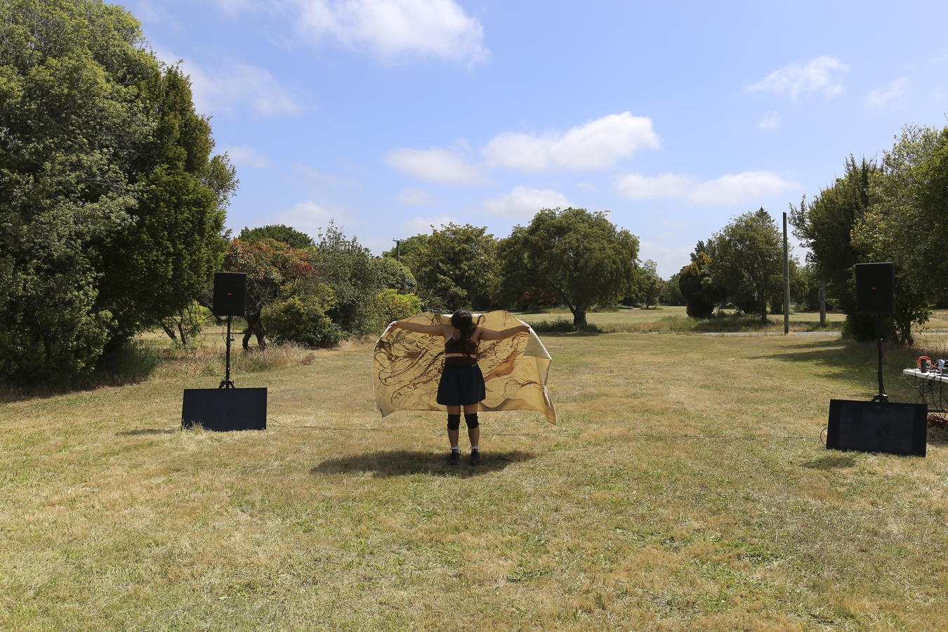 Ella standing on the plot of grass where her family home once stood. a canvas draped over her shoulders, arms spread like a butterfly.