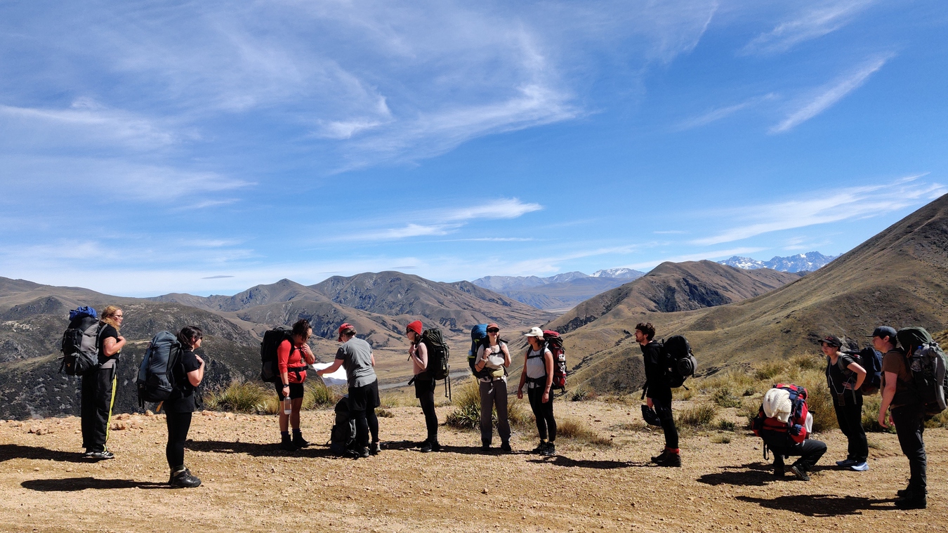 A group of us with hiking gear and packs still on stand at a plateau, Chloe pulls out a topographical map, a mountain range with light dustings of snow create a striking backdrop in the distance