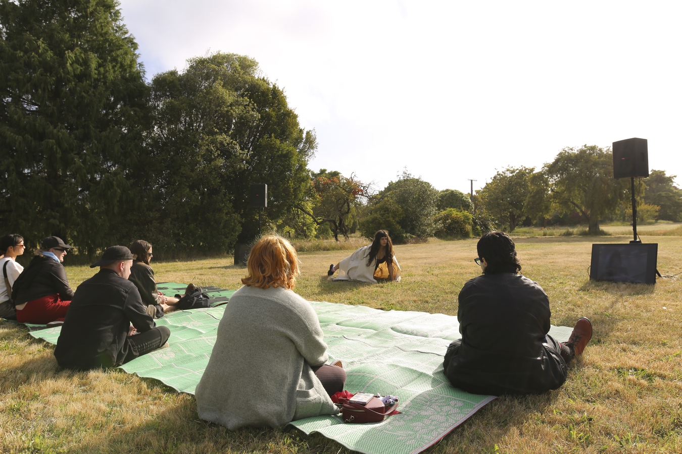 The audience sitting on mats watching Ella perform. Ella is lunging forward with her hands planted on the ground, the sun glowing behind her