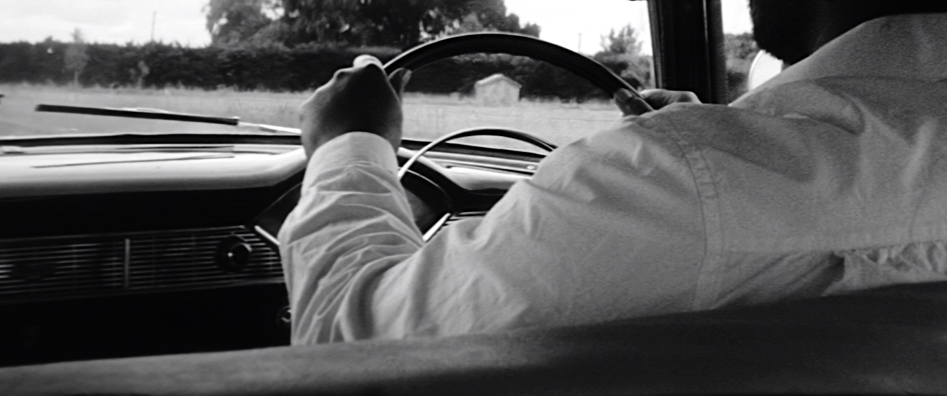 A black and white film still shows the back of a man driving a vintage Chevy Bel Air car. His hands are holding the steering wheel