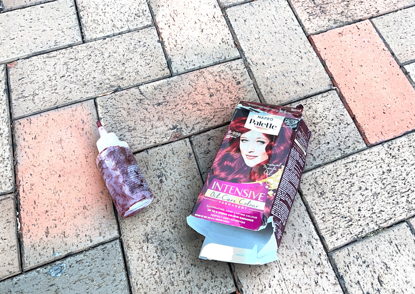 A Palette hair dye box that has been opened on the ground next to a hair dye container stained by hair dye. The ground is interconnected red and grey rectangular paving stones.