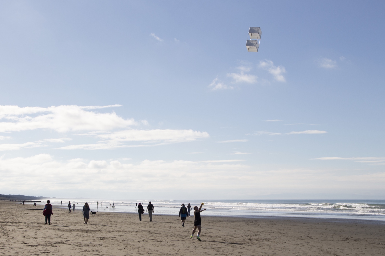 sandy beach with a box kite flying, a small crowd and an adolescent running the kite, blue sky