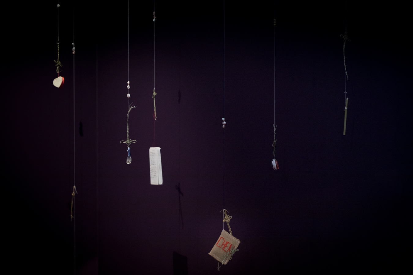 Seven objects including a hand made book cover, small glass bottle, a ceramic dish and hand written recipe on paper are suspended from the ceiling on knots made of harakeke